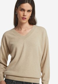 Beige V-Ausschnitt Pullover aus weichem Strickmaterial. Hat lange Ärmel und einen gerippten Saum. Das Modell posiert vor einem neutralen Hintergrund.