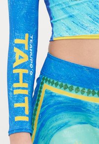 Blå långärmad topp med gult "TAHITI"-tryck, strukturerad yta, kombinerad med blå mönstrade byxor med gröna och gula geometriska accenter.
