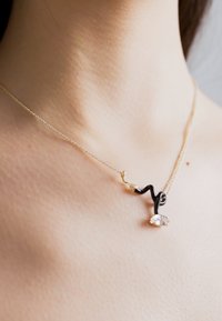 Culoare neagră și aurie, această colier are un design răsucit și o piatră ovală clară, așezat pe pielea goală. O lanț subțire completează aspectul minimalist.