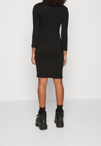 Černé dlouhé bodycon šaty s přiléhavým střihem. Nosené s černými robustními botami a ponožkami, texturovaná látka, žádné viditelné vzory.