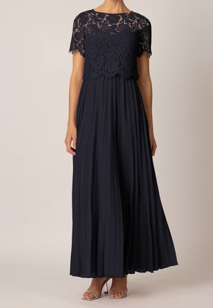 Femme portant une robe bleu marine avec un haut en dentelle à manches courtes et une longue jupe plissée, associée à des talons ouverts argentés.