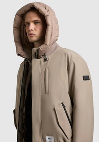 Beige színű pufajka, nagy, steppelt kapucnival. A dizájn tartalmaz egy cipzáras zsebet, egyenes szabást és fekete címkét az ujjon.