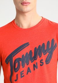 Czerwony bawełniany T-shirt z wyraźnym niebieskim logo "Tommy Jeans" w kursywie. Okrągły dekolt, luźny krój, gładka tekstura.