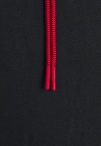 Deux extrémités de lacets rouges rapprochées sur un fond de tissu noir texturé, montrant le tissage détaillé et les embouts en plastique.