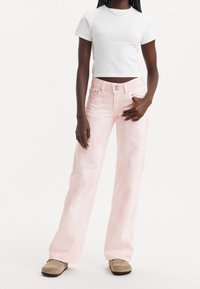 Femme portant un t-shirt blanc court, un jean large rose clair et des chaussures en suède beige devant un fond blanc.