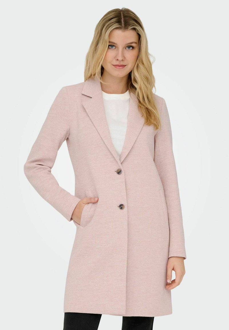 Ženska oblečena v svetlo roza dolg plašč z dvema gumboma in žepoma, pod njim bela majica in črne hlače, stoji ob preprosti ozadju.