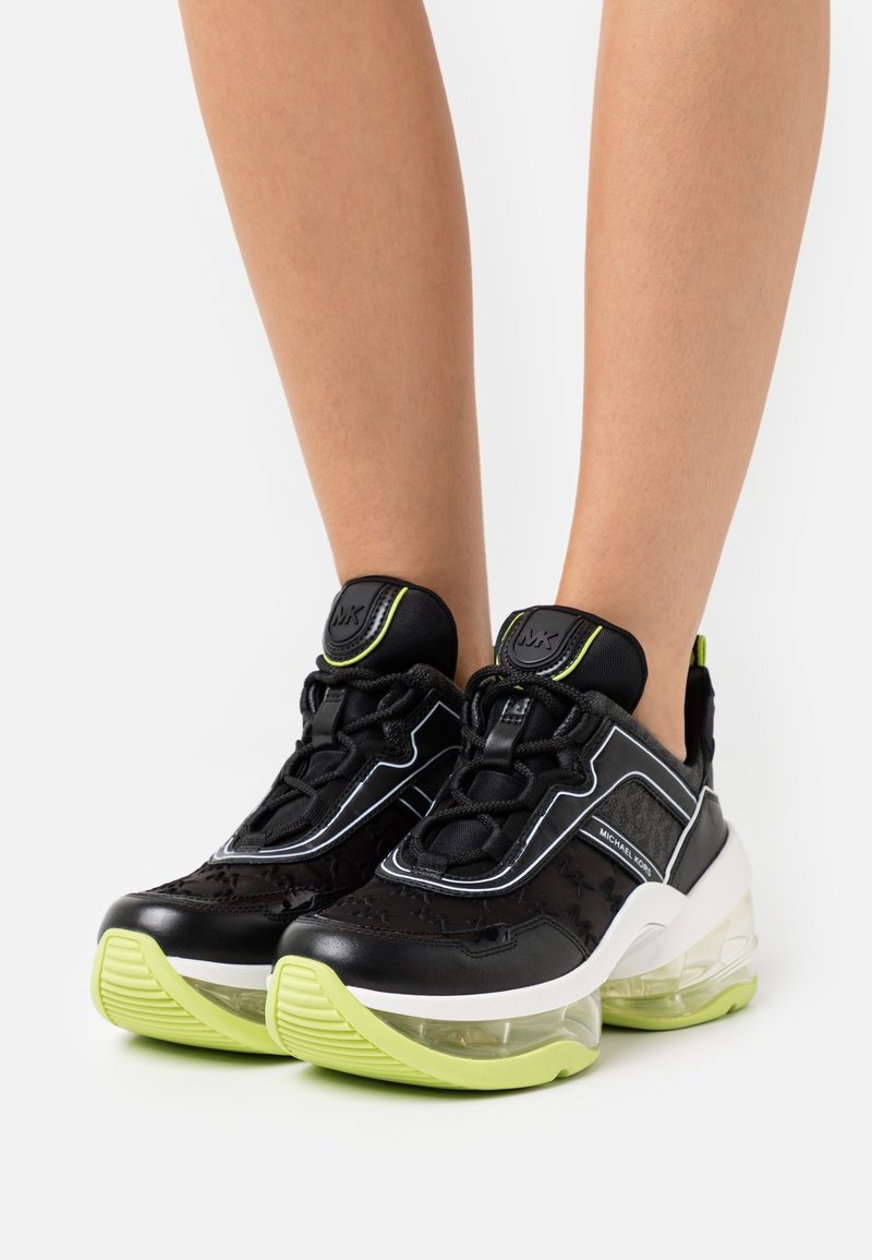 Baskets Michael Kors noires et vert citron avec des semelles épaisses transparentes portées sur des jambes nues devant un fond uni.