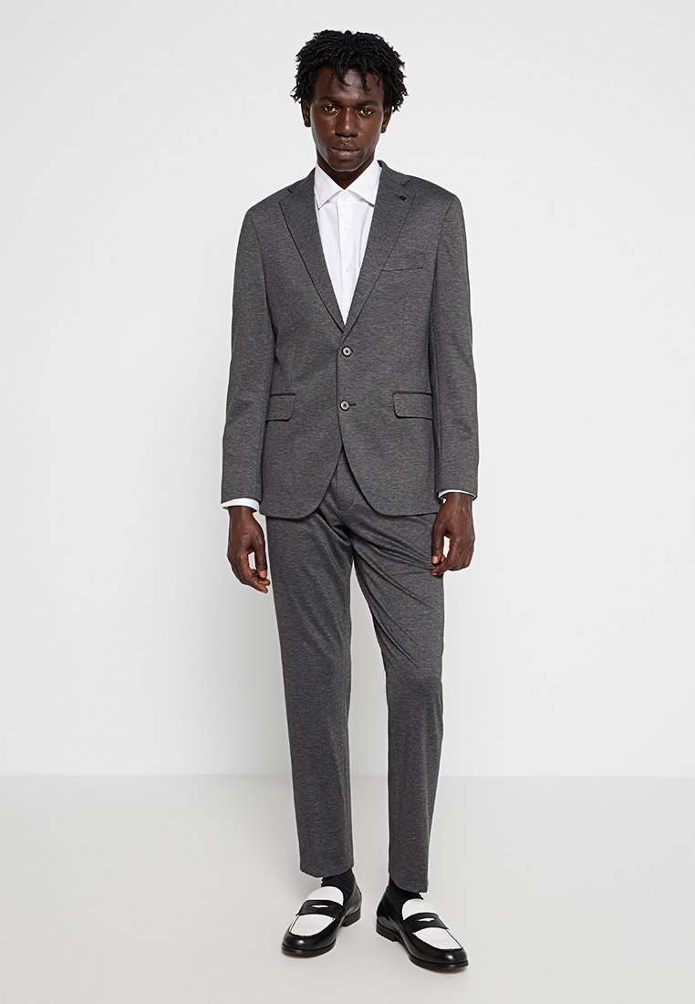 Traje gris a medida con chaqueta de dos botones y pantalones de frente plano. Combinado con una camisa blanca de botones y zapatos en blanco y negro.