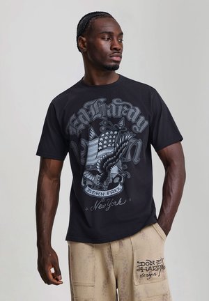 Hombre con camiseta negra gráfica con diseño de águila y bandera americana y pantalones beige con texto "Don Ed Hardy designs" en el bolsillo.