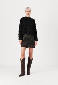 Giacca nera dalla texture lavorata con design a ruches, abbinata a una mini gonna di pelle scura. Stivali da cowboy marroni al ginocchio con punte appuntite.