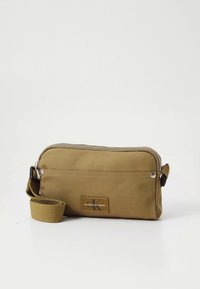 Borsa in tessuto verde oliva con chiusura zip, tasca frontale e patch con marchio. Include una tracolla removibile per versatilità. Forma rettangolare.