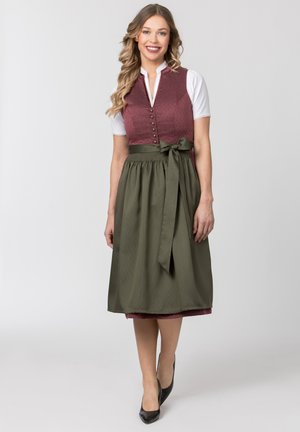Frau mit langen lockigen Haaren, die ein bordeauxfarbenes ärmelloses Kleid, eine weiße Bluse, eine olivgrüne Schürze, am Gürtel gebunden, und schwarze Absätze trägt, lächelt.
