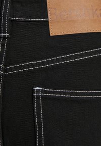 Svart denimtyg med kontrasterande vit söm, försett med en läderetikett som står "Bershka." Inkluderar en bakfickadetajl.