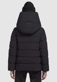 Henkilö pukeutuneena mustaan toppatakkiin, jossa on suuri huppu, ja mustaan neulottuun pipoon, selin vaaleaa yksiväristä taustaa vasten.