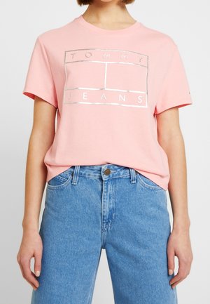 Persoon draagt een roze Tommy Jeans T-shirt met zilveren logoprint, gecombineerd met een high-waisted blauwe spijkerbroek.
