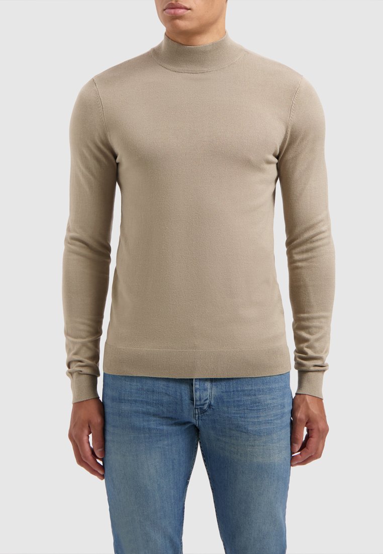 Beige gebreide trui met een hoge hals, geribbelde manchetten en zoom, gecombineerd met lichtblauwe jeans. Soepele textuur en een getailleerd ontwerp.