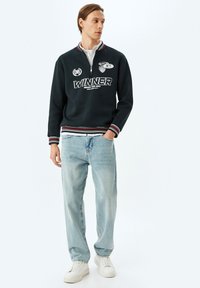 Sudadera de color marino con cuello con cremallera, puños blancos y texto bordado. Jeans de mezclilla de color azul claro y zapatillas blancas completan el atuendo. Diseño casual.