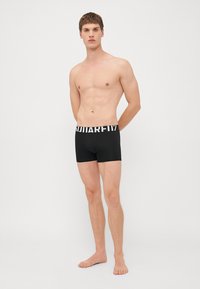 Fekete boxer alsónadrág fehér márkázott derékpánttal. Simább, rugalmas anyagból készült. Testhez simuló dizájn és térd közepéig érő hosszúság.