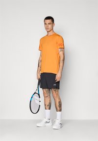 Oranžinė marškinė su raudonu logotipu, juodos šortai, balti sportbačiai, laikantis teniso raketę su mėlynais akcentais ir matomais tatuiruotėmis ant rankų ir kojų.