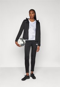 Svart zip-up jacka, vit mönstrad skjorta, svarta sportbyxor med sidostripes och svarta sportskor; håller en svart och vit fotboll.