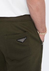 Olivgrüne Jogginghosen aus Baumwolle mit einem gerippten Bund, einer Gesäßtasche mit silbernem "OMBRE"-Logo und einem Reißverschlussdetail an der Tasche.