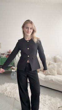 Ung kvinna med blont hår som bär en svart knäppt kavaj och svarta byxor, stående i ett mysigt vardagsrum med neutral inredning.