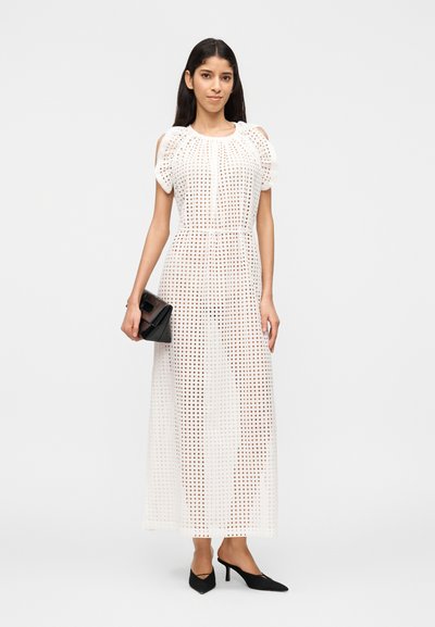 Γυναίκα που φοράει λευκό μακρύ φόρεμα με τρύπες, κρατώντας μαύρο τσαντάκι χειρός, και φορώντας μαύρες μυτερές γόβες, στέκεται σε λευκό φόντο.