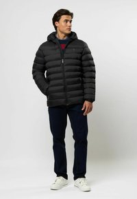 Mann in schwarzer Steppjacke mit Kapuze, dunklen Jeans und weißen Sneakers, der steht und zur Seite schaut, vor einem schlichten Hintergrund.