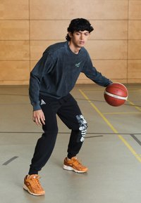 Black sweatpants with white graphic print, gray long-sleeve shirt with green logo, and orange sneakers, dribbling a basketball indoors.
