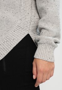 Hellgrauer Pullover mit strukturiertem Strick, ausgestattet mit einem gerippten Saum und Ärmeln sowie dezente schwarze Sprenkel im gesamten Design, kombiniert mit schwarzen Hosen.