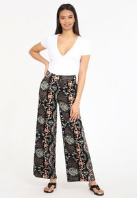 Haut blanc à manches courtes avec un décolleté en V profond, associé à un pantalon large à motifs noir, vert et corail. Sandales avec une boucle pour les orteils.