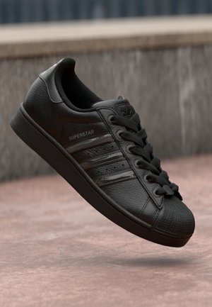 Baskets Adidas Superstar noires en cuir texturé, bout coquille, lacets noirs et bandes de la marque, montrées flottant au-dessus d’un sol pavé rosé.