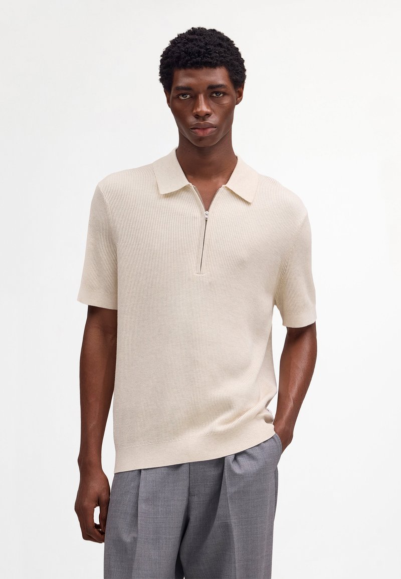 Giovane uomo che indossa una polo lavorata a maglia beige a maniche corte con cerniera e pantaloni grigi plissettati, in piedi davanti a uno sfondo bianco uniforme.