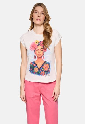 Mujer con cabello largo y rubio, lleva una camiseta blanca con un retrato floral colorido y pantalones rosas, de pie frente a un fondo liso.