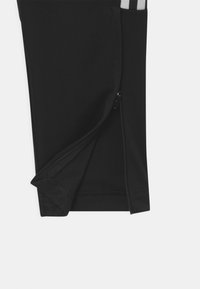 Zwarte sportbroek met een ritssluiting aan de pijp, voorzien van een gladde textuur en versterkte stiksels langs de zoom. Geaccentueerd met grijze strepen.