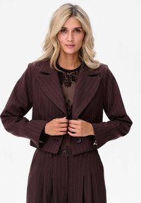 Brązowa marynarka w prążki wykonana z teksturowanej tkaniny, z szerokim klapą i dwoma guzikami, noszona na przezroczystej kwiatowej bluzce.