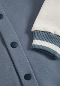 Blauwe trui detail met marineblauwe knopen, gestructureerde stof en een witte geribbelde manchet met blauw-witte strepen. Zachte textuur, casual ontwerp.