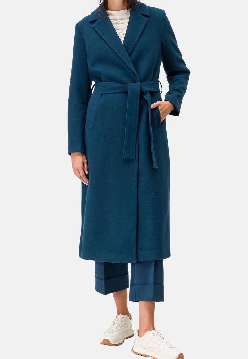 Long manteau en laine bleu sarcelle avec ceinture, col à revers et poches, associé à un pantalon bleu court et des baskets beige. Texture douce.