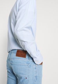 Camisa de manga larga azul claro combinada con jeans de denim de lavado claro, que presentan un parche de cuero marrón de Tommy Hilfiger en la parte posterior.
