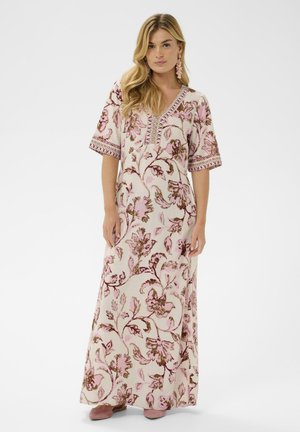 Femme portant une longue robe à motifs floraux roses et beiges avec des manches courtes, ainsi que des boucles d'oreilles et des chaussures roses assorties.