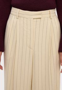 Cremefarbene gestreifte Hose mit Falten, ausgestattet mit einem taillierten Bund und Seiten Taschen, kombiniert mit einem burgunderfarbenen Pullover.