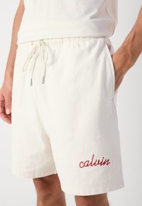 Valkoiset puuvillashortsit, joissa on elastinen vyötärö ja naru. Vasemmassa lahkeessa on brodeerattu brändinimi "calvin" punaisena.