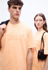 Νέος άνδρας και γυναίκα φορούν ταιριαστά ροδακινί T-shirts «Tommy Jeans», ο άνδρας κρατάει ένα σακάκι στον ώμο του, η γυναίκα κρατά μια μαύρη τσάντα.