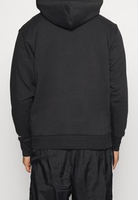 Sweat à capuche noir avec une coupe décontractée, une capuche réglable et des poignets côtelés. Présente une texture lisse et des coutures le long des bords.