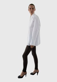 Camicia bianca con bottoni e maniche lunghe, abbinata a leggings neri e scarpe con tacco, su uno sfondo neutro.