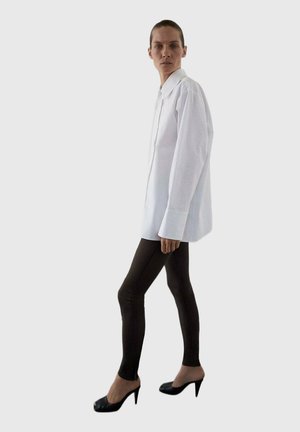 Camisa blanca de botón con mangas largas, combinada con leggings negros y zapatos de tacón alto, contra un fondo sencillo.