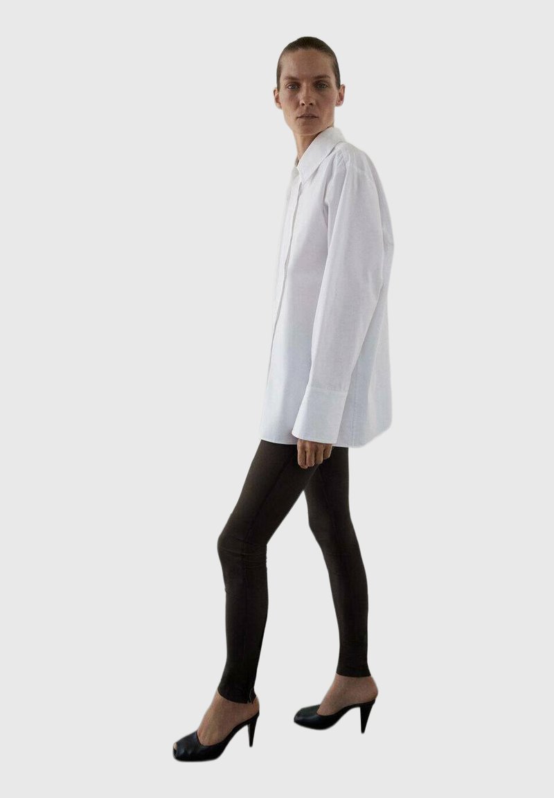 Camicia bianca con bottoni e maniche lunghe, abbinata a leggings neri e scarpe con tacco, su uno sfondo neutro.