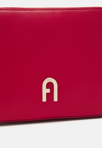 Rød læderpung med et guldfarvet metallogo i form af et "a". Glat tekstur med rene kanter og minimalistisk design.