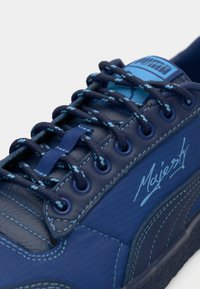 Navy blue sneaker with textured leather and nylon materials, featuring contrasting light blue laces and stitching, and a logo on the side.