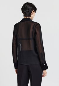 Camicia nera trasparente con maniche lunghe, scollo con colletto e polsini con bottoni. Texture notevole con sottili linee verticali. Tessuto a strati sottili.